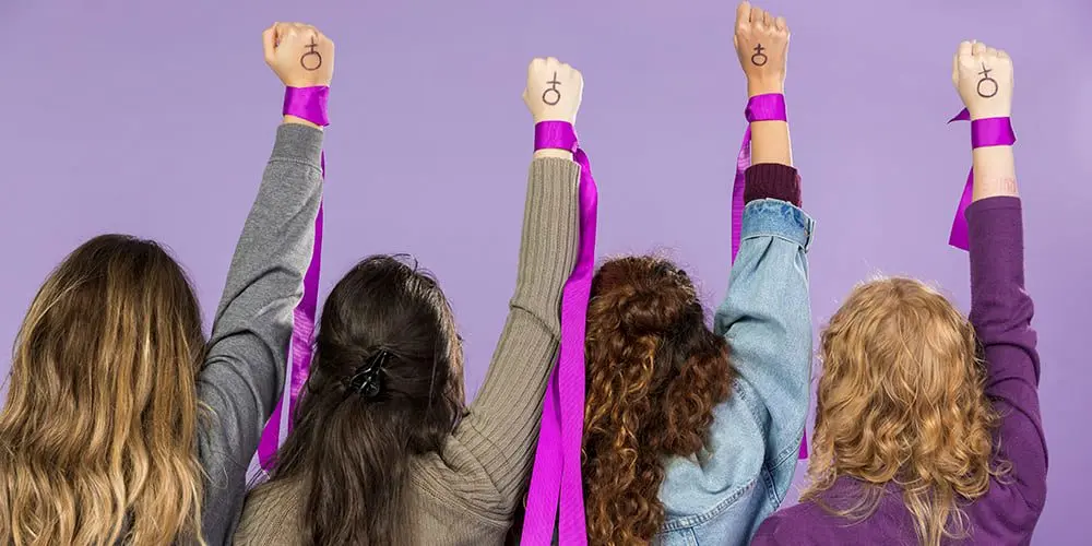 Mujeres alzando su brazo derecho con un muñequera de color morado, en el puño de la mano tienen el genero femenino, en un fondo morado.